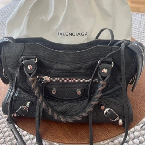 ÄKTA svart Balenziaga väska i st small - Säljer min snygga svarta handväska från Balenciaga i mjukt skinn med silverfärgade metalldetaljer, flätat handtag och dragkedja framtill. I st liten. En metalldetalj är av där fram som syns på bild 4. Påse axelband och kvitto följa med. Pris kan diskuteras  