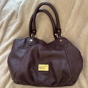 Mörkbrun/lila handväska Marc by Marc Jacobs - Säljer en snygg mörkbrun/lila handväska från Marc by Marc Jacobs i äkta skinn. Väskan har två rejäla handtag, guldfärgade metalldetaljer och en klassisk logoplatta framtill. Perfekt storlek för att få plats med allt du behöver. Jättefint skick. Kan mötas upp i Stockholm och frakta. 