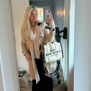 Beige Tote Bag från Marc Jacobs - Säljer en beige Tote Bag från Marc Jacobs med svart text och bred svart axelrem. Väskan är fyrkantig med två handtag och avtagbar axelrem. Tillverkad i kraftig bomullscanvas och har tydlig logga framtill. Perfekt för dig som vill ha en trendig och rymlig väska. 😍Säljer pga att jag vill köpa en ny innan färg. Den är sparsamt använd och är som ny. Nypris 4000kr, säljer för 3000kr. 🌸🥰