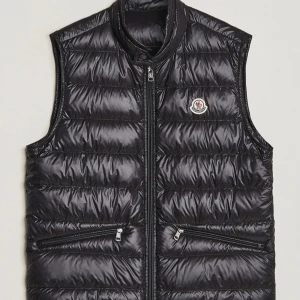 Svart Moncler gui väst - Snygg svart dunväst från Moncler med quiltad design och glansig finish. Västen har hög krage, två dragkedjefickor framtill och klassisk Moncler-logga på bröstet. Perfekt att slänga över en hoodie eller tröja för extra värme.