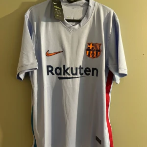 FC Barcelona Nike fotbollströja ljusblå - Snygg ljusblå FC Barcelona fotbollströja från Nike med klubbmärke och orange swoosh på bröstet. Rakuten-logga framtill och Unicef-tryck bak. Tillverkad i lätt och ventilerande polyester, perfekt för match eller träning. Klassisk passform med korta ärmar.