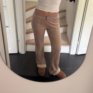 Beige velourbyxor Juicy Couture S - Mysiga beige mjukisbyxor från Juicy Couture i velour med raka ben, hög midja och snörning i midjan. Byxorna har stora fickor fram och bak med knappdetaljer. Perfekta för en chill och trendig look.