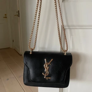 Svart väska från Saint Laurent - Säljer en svart axelväska från Saint Laurent med kedjerem i guld och ikoniskt YSL-emblem framtill. Väskan är i skinn och har en stilren, klassisk design som passar till många outfits. 