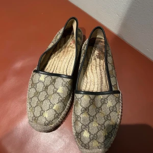 Gucci espadrillos med GG-mönster - Snygga espadrillos från Gucci med klassiskt GG-mönster i beige och guld, samt svarta detaljer runt öppningen. Skorna har flätad sula och är perfekta för dig som vill ha en lyxig och trendig look. Materialet är canvas med inslag av skinn. Inget synligt slitage på utsidan men insidan har loggan försvunnit. 