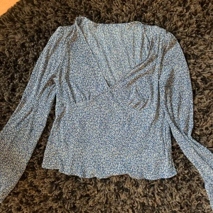 Blå blommig omlottblus med v-ringning - Supersöt blå blus med små vita blommor, omlottmodell och v-ringning. Långa ärmar och lätt vågig nederkant ger en romantisk vibe. Perfekt för dig som gillar en feminin och trendig stil.