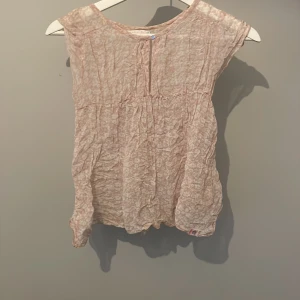 Ljusrosa blommig blus i tunn  - Supersöt ljusrosa blus med småblommigt mönster och lätt transparent tyg. Modellen är ärmlös med rund halsringning och har en luftig, flowy passform. Perfekt för varma dagar när du vill ha något lätt och romantiskt.