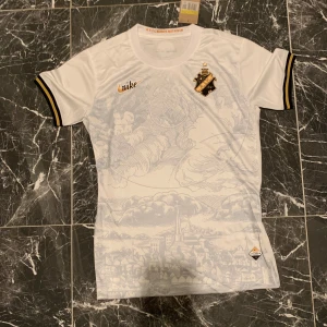 AIK vit limeted edition Nike 2023 - AIK:s vita med guldfärgade detaljer och klubbmärke på bröstet. Tryckt motiv av Stockholm på framsidan och svarta/gula ränder på ärmsluten. Tröjan är i lätt och ventilerande material, perfekt för match eller träning.