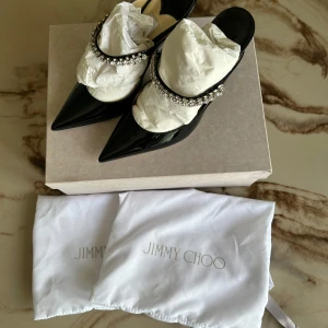 Svarta pumps med strass från Jimmy Choo - Supersnygga svarta pumps från Jimmy Choo i lackat skinn med spetsig tå och hög klack. Skorna har en elegant rem dekorerad med glittrande strass som ger extra lyxig känsla. Perfekta för dig som vill sticka ut med stil.