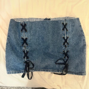 Blå jeanskjol med snörning - Säljer en blå jeanskjol med rå kant upptill och snygga svarta snörningsdetaljer framtill. Kjolen är kort och har dragkedja bak. 