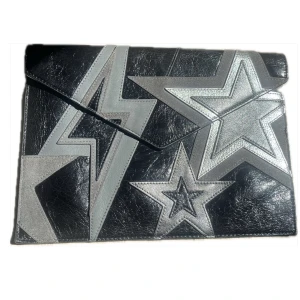 Svart och silver kuvertväska med stjärnor - Kuvertväska i svart och silver skinn med stora stjärn- och blixtdetaljer på framsidan. Väskan har ett coolt, grafiskt mönster och stängs med ett lock. Perfekt för dig som vill sticka ut med accessoarer. Från rebecca minkoff, nypris 1895:-