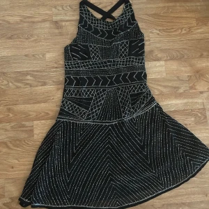 Svart kortklänning Glamorous S - Svart kortklänning från Glamorous i storlek S. Klänningen har silvriga glittriga mönster, korsade axelband i ryggen och en figurnära överdel med utsvängd kjol. Perfekt för dig som vill sticka ut med coola detaljer och snygg passform.