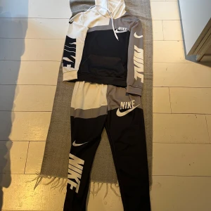 Nike mjukisbyxor/tröja  i svart, grå och vit - Snygga mjukisbyxor från Nike i färgblockad design med svart, grå och vit. Byxorna har resår i midjan, smal passform och stora Nike-loggor på benet. Perfekta för chill eller träning och tillverkade i mjuk polyesterblandning. Kan även passa till st s 