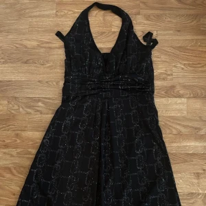 Svart viscosa glittrig långklänning med halterneck  - Svart långklänning med halterneck och glittrigt mönster. Djup v-ringning framtill och markerad midja. Klänningen har ett snyggt fall och är perfekt för dig som vill sticka ut med lite extra glitter.