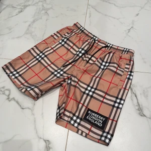 Rutig Burberry shorts med snörning - Snygga shorts från Burberry med klassiskt rutigt mönster i beige, svart, vitt och rött. De har elastisk midja med svart snörning och en patch med Burberry London England på benet. Perfekta för sommaren och ger en clean streetstyle-vibe.