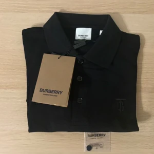 Svart pikétröja från Burberry - Klassisk svart pikétröja från Burberry med diskret broderad logga på bröstet. Tröjan har krage, knäppning med tre knappar och är tillverkad i mjuk bomull. Perfekt för en stilren och clean look.