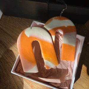 Stora orangevita bågformade örhängen - Unika, handgjorda örhängen i stor bågform med coolt randigt mönster i orange och vitt. Statement-accessoar som verkligen sticker ut och ger en färgklick till din look. Lätta att bära och perfekta för dig som gillar att synas.