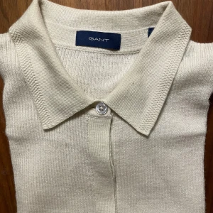 Beige stickad pikétröja från Gant - Snygg beige stickad pikétröja från Gant i storlek XS. Tröjan har klassisk krage, kort ärm och knäppning framtill med en knapp. Perfekt för dig som gillar en clean och stilren look. Materialet är mjukt och ger en skön känsla.