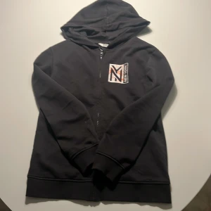 Svart hoodie med New York-tryck H&M - Svart hoodie från H&M med dragkedja och huva. På bröstet finns ett tryck med texten 'New York' och ett stort M i vitt, orange och svart. Tröjan är i mjuk bomull och har långa ärmar och ribbade muddar.
