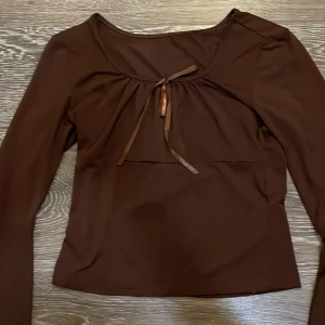 Brun långärmad topp med rosett - Mörkbrun långärmad topp med söt rosettdetalj framtill och rundad urringning. Toppens material är stretchigt och har en slät yta, vilket gör den bekväm och smickrande. Perfekt för dig som gillar en enkel men ändå trendig look. Däremot på högra armen så är det två små hål men de syns knappt, den passar på XS och S