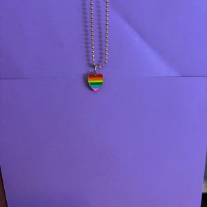 Halsband med regnbågshjärta - Snyggt halsband med kulkedja och ett hjärtformat hänge i regnbågens färger. Perfekt accessoar för att visa din pride eller bara addera lite färg till din stil. Hjärtat har tydliga ränder i rött, orange, gult, grönt, blått och rosa.