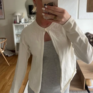 Vit kofta med knapp i halsen - Säljer en stilren vit kofta med rund halsringning och en enkel knapp högst upp. Koftan är långärmad och har en mjuk, tunn känsla som passar perfekt över ett linne eller t-shirt. Enkel och clean design som funkar till många outfits.