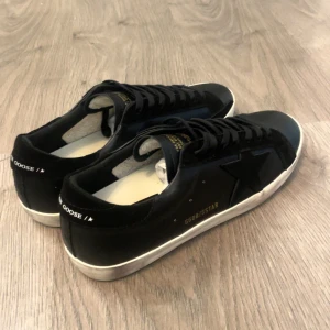 Golden Goose Super-Star  - Svarta Golden Goose Super-Star sneakers med vit sula och svart stjärndetalj på sidan. Skorna har snörning, rund tå och är tillverkade i skinn och mocka. Det är storlek 44 och den är 27 cm i insidan. Ställ frågor vid fundering. Box medföljer ej. 