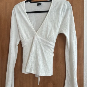 Tie top Gina tricot - Säljer en vit omlottopp från Gina tricot med lång ärm och v-ringning. Toppen har en snygg struktur i tyget och knytband i midjan som ger en fin passform. Perfekt att styla med jeans eller kjol för en fräsch look. Den passar både L och M jag har själv M/S och den passa mig. Den har aldrig haft på mig den så nu bestämde jag mig att sälja den!!