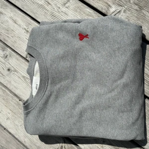Grå sweatshirt från Ami Paris - Snygg grå sweatshirt från Ami Paris med klassisk rund hals och röd broderad logga med hjärta på bröstet. Tröjan är i mjuk bomull och har ribbade muddar vid ärmslut och nederkant. Perfekt för en chill och stilren look.