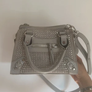 Grå handväska med nitar och dragkedja - Snygg grå handväska med silvriga nitar och detaljer. Väskan har både handtag och axelrem, samt en dragkedja framtill. Perfekt för dig som vill ha en cool och trendig accessoar med lite edge. Helt oanvänd.