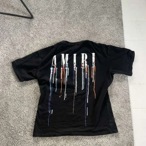 Svart Amiri t-shirt med färgstänk - Svart t-shirt från Amiri med stort tryck på ryggen och mindre tryck på bröstet. Trycket består av vita bokstäver och färgstänk i blått, orange och vitt som rinner ner. Klassisk passform och korta ärmar, tillverkad i mjuk bomull.