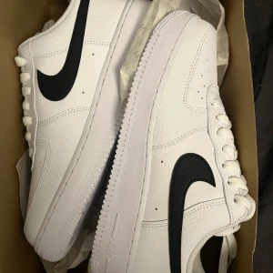 Nike Air Force 1 vit/svart strl 40 - Nike Air Force 1 i klassisk vit med svart swoosh. Skorna har en chunky sula, perforerad tå och är gjorda i skinn med snörning. Perfekt för dig som vill ha en clean och tidlös sneaker med streetkänsla. Dom är aldrig ens provade, lådan medföljer. Nypris ca 1000kr