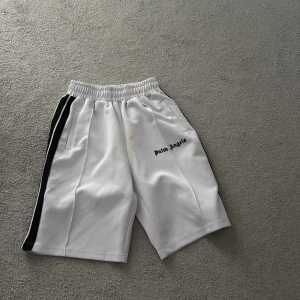 Palm Angels vita shorts med svarta ränder - Snygga vita shorts från Palm Angels med svarta ränder längs sidorna och logga tryckt på ena benet. De har elastisk midja och sportig vibe, perfekta för en chill och trendig look.