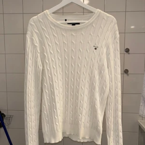 Vit kabelstickad tröja från Gant - Vit kabelstickad tröja från Gant med rund halsringning och diskret logga på bröstet. Tröjan har lång ärm och klassisk, stilren look som funkar till allt. Perfekt för dig som gillar preppy vibes och enkel stil. Den sitter som S.