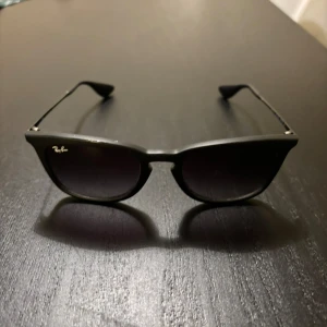 Svarta Ray-Ban solglasögon - Klassiska svarta Ray-Ban solglasögon med fyrkantig form och mörka glas. Snygg och tidlös design med diskret Ray-Ban-logga på ena linsen. Bågarna är i plast och skalmarna är tunna och svarta. Perfekta för en cool och stilren look.