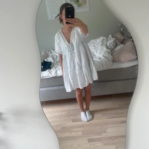 Vit kort klänning med spetsdetaljer - Supersöt vit kortklänning i luftig bomull med v-ringning och fina virkade spetsdetaljer längs halsringningen och ärmsluten. Klänningen har en lös passform och korta ärmar, perfekt för varma dagar. Ger en fräsch och somrig vibe!