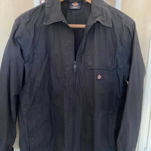 Svart jacka från Dickies - Säljer en svart jacka från Dickies med dragkedja och en bröstficka med knappdetalj. Jackan har en klassisk krage och långa ärmar. Perfekt för en stilren look.