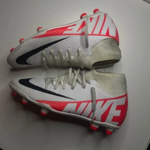 Nike mercurial superfly 9 - Snygga fotbollsskor från Nike med en iögonfallande röd och vit design. Skorna har snörning och en svart Nike-logga på sidan. Perfekta för fotbollsplanen med sina röda dobbar för optimalt grepp.