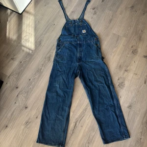 Carhartt Blå denim overall - Säljer en klassisk blå denim overall från carhartt med justerbara axelband och flera praktiska fickor. Perfekt för en avslappnad och trendig look. Overallen har en rak passform och är tillverkad i slitstarkt jeansmaterial. Knappt använda. Då de inte direkt e min still. Priset är inte hugget i sten. Hör av dig angående mer frågor. Jag som är 1,76 passade i overallen 