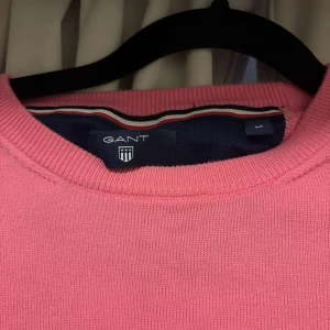 Rosa tröja från GANT - Säljer en snygg rosa tröja från GANT med rund halsringning. Tröjan har långa ärmar och ett diskret broderat märke på bröstet. Perfekt för en stilren look. Storlek M