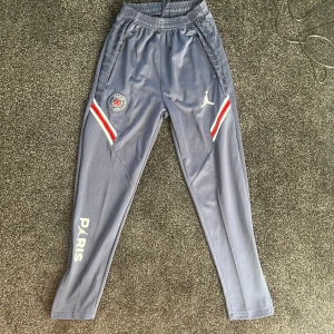 Grå träningsbyxor från Jordan x PSG - Snygga grå träningsbyxor från Jordan x PSG med elastisk midja och snörning. Byxorna har vita och röda detaljer samt logotyper på benen. Perfekta för sport och vardag.