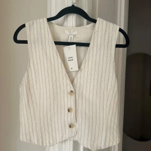 Vit randig väst från H&M - Snygg vit väst i linneblandning från H&M med smala svarta ränder. Västen har en klassisk design med knappar framtill och en stilren look. Perfekt för att lägga till en touch av elegans till din outfit. I butik kostar den 199, osäker om den ens finns kvar. Mitt pris 150