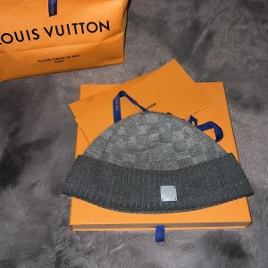 Grå mössa från Louis Vuitton - Snygg grå mössa från Louis Vuitton som är köpt i butiken på Östermalm. Diskret mönster och en liten logotyp framtill. Mössan har en ribbad kant som ger en stilren look. Låda, box, kvitto och påse finns.