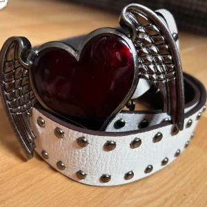 Vintagebälte med nitar! - Vintagebälte i vitt läder med nitar och bältesspänne med hjärta med vingar. L: 110 cm (78-99 cm innersta till yttersta hålet). B: 4 cm. Oanvänt.