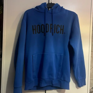 Blå hoodie från Hoodrich - Säljer en snygg blå hoodie från Hoodrich med svart logotyp på bröstet. Tröjan har en klassisk känguruficka och justerbar huva med dragsko. 