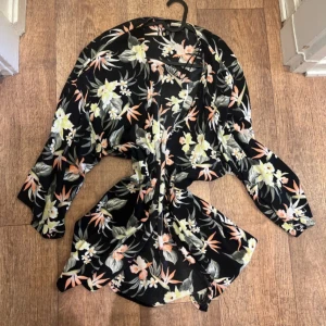 kimono  - Snygg kimono med blommigt mönster i svart, orange och vitt. Den har en lös passform och trekvartsärmar, perfekt för en avslappnad stil. Knytband i midjan för en justerbar passform. Snabb leverans 📦 