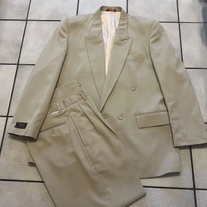 Beige kostym från Lawell - Elegante beige kostym från Lawell med kavaj och matchande byxor. Kavajen har klassiska slag och två knappar framtill. Byxorna har en stilren design med hällor för bälte. Perfekt för formella tillfällen. kavaj längd 78 cm, omkrets under armen 114 cm, ärmlängd 124 cm. Byxa längd 108,5 cm, midja omkrets byxor 78 cm, 
