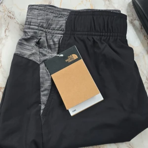 Svarta träningsshorts från The North Face - Snygga svarta träningsshorts från The North Face med grått mönster på sidorna. De har en elastisk midja för extra komfort och är perfekta för träning. Shortsen är tillverkade i ett lätt och andningsbart material.Helt oanvända. Ny pris €50 