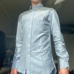 Ljusblå skjorta från Ralph Lauren - Säljer en stilren ljusblå skjorta från Ralph Lauren i slim fit. Skjortan har långa ärmar och knappar framtill samt en broderad logotyp på bröstet. Perfekt för en elegant look. Jag är 175 och den satt som en smäck så 170-178 är nog en bra passform!
