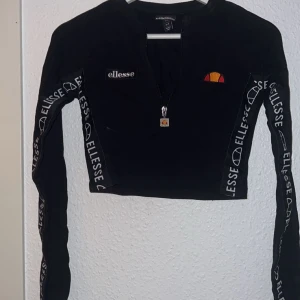 Svart croppad tröja från Ellesse - Snygg svart croppad tröja från Ellesse med dragkedja och logga på bröstet. Ärmarna har Ellesse-tryck längs sidorna. Detta är en sport tröja men funkar super bra till vardags tröja också. Lite mer åt magtröja hållet. Nypriset: 600. 