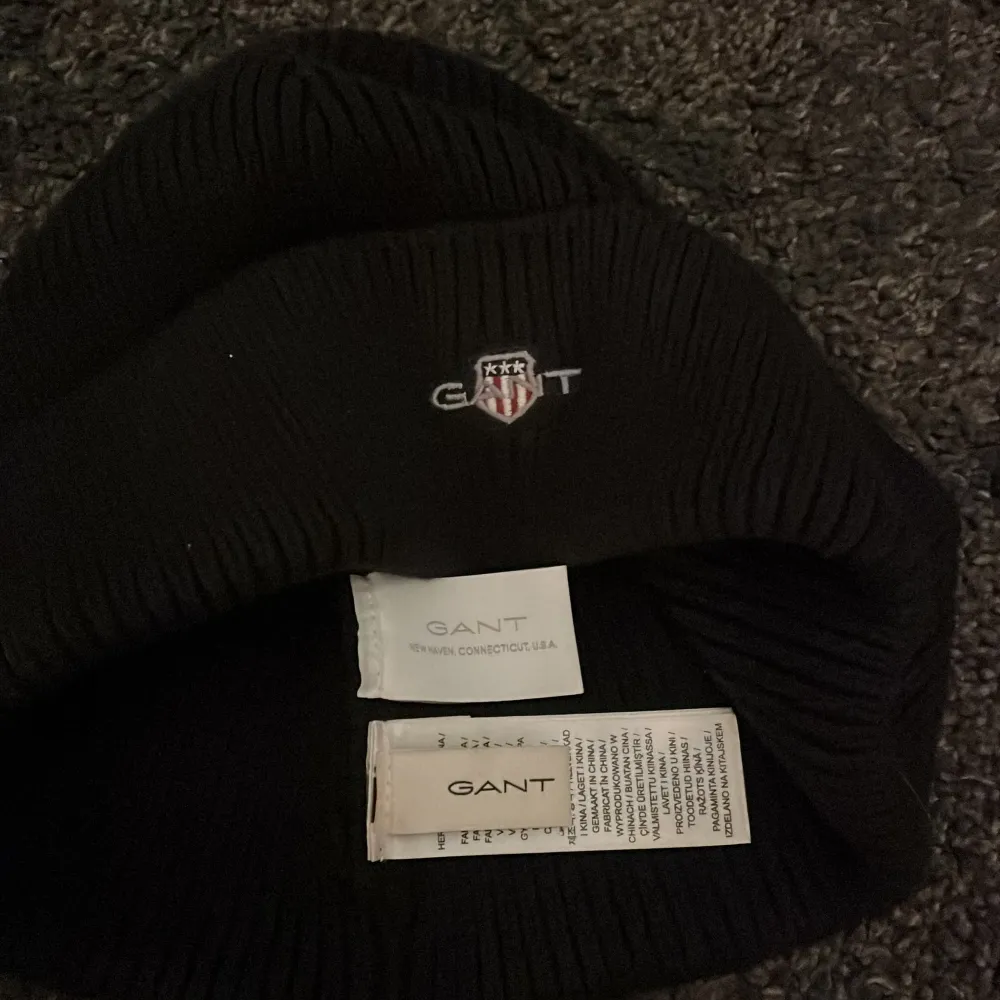 Snygg svart mössa från GANT med ribbad design och broderad logotyp framtill. Perfekt för att hålla värmen under kyliga dagar.. Asusteet.